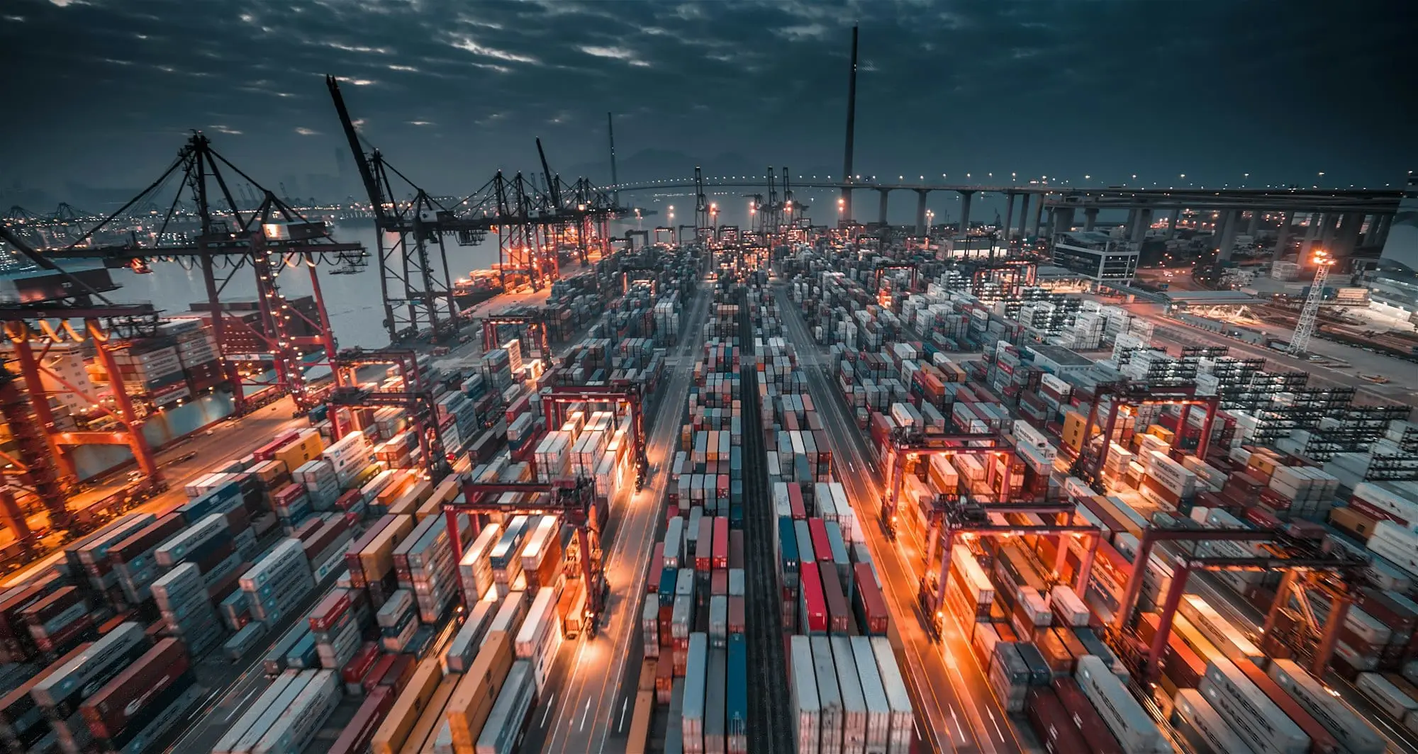 Container port in Hong Kong at dusk, with stacked shipping containers and gantry cranes lit up