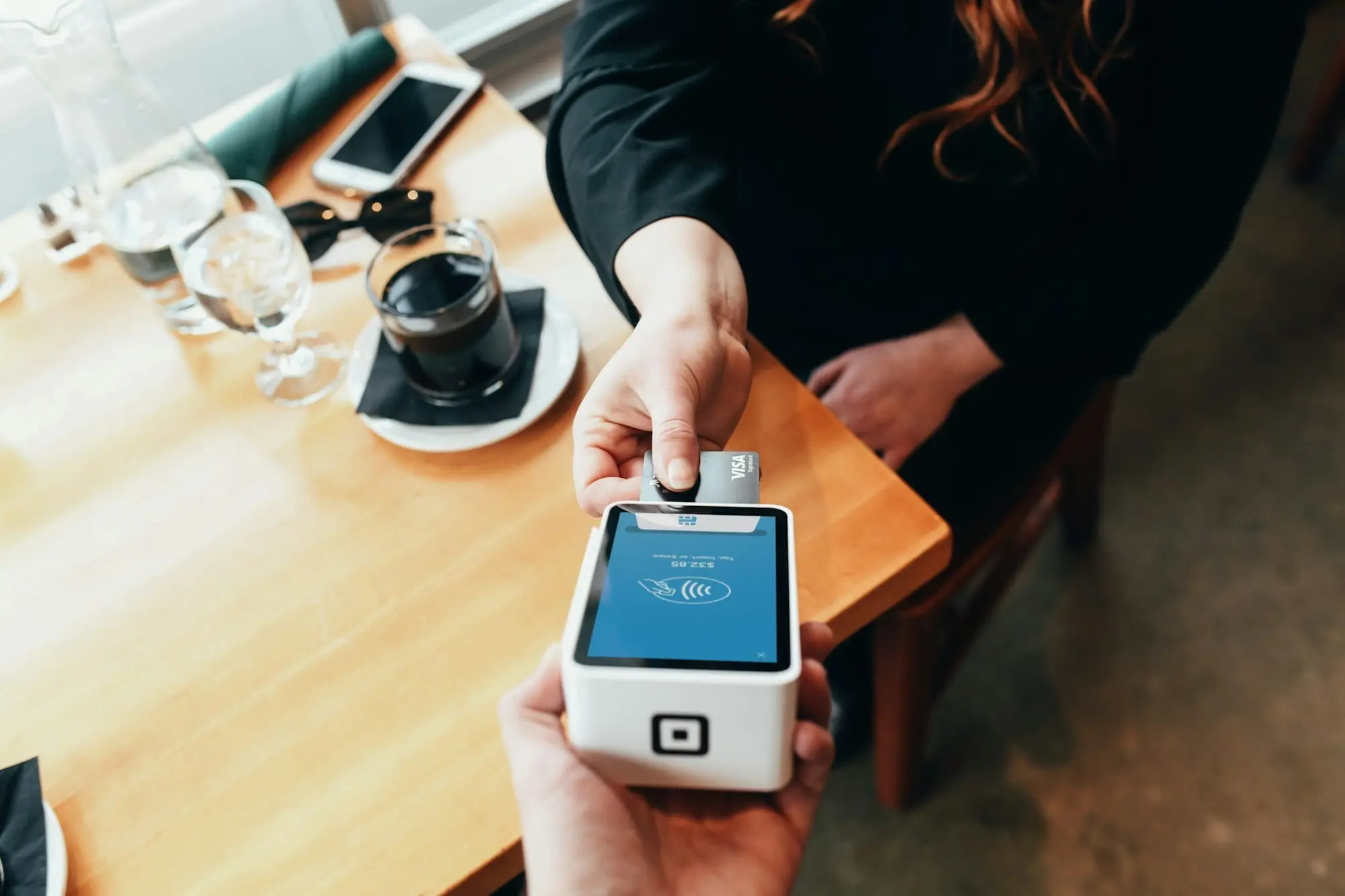 A customer taps a card on a handheld payment terminal held out across a cafe table
