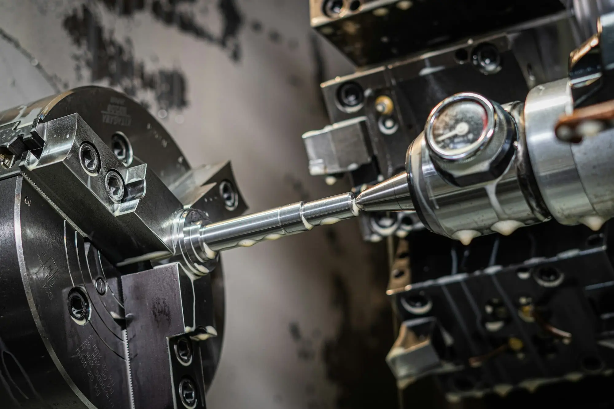 A steel shaft being turned on a CNC lathe, clamped in a three-jaw chuck and supported by a live centre