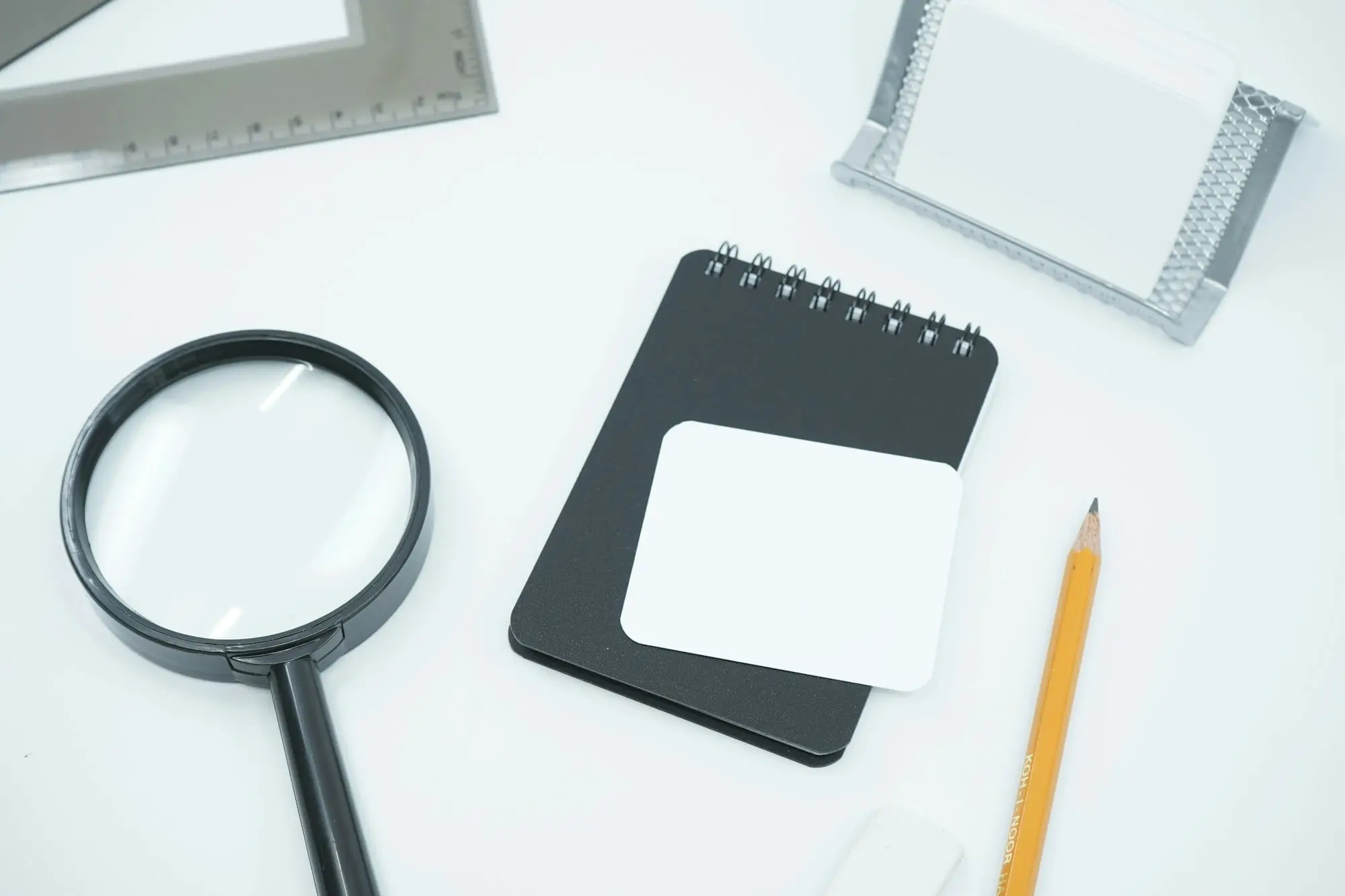 A magnifying glass, a black notepad and a pencil laid out on a white desk
