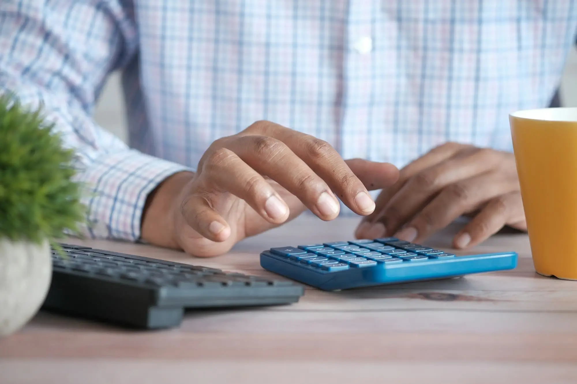 Hands working a calculator at a desk next to a keyboard and a coffee mug